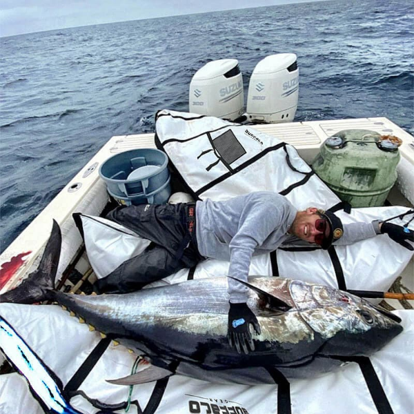 A large tuna fish lies on white and black gear bags inside a boat deck, with a person lying beside it. Two Suzuki outboard motors are visible in the background, along with a blue bucket and a green gas canister, with open ocean water behind them.