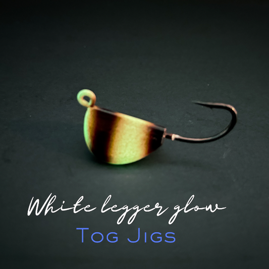 Fishing lure with gradient color on a dark background, labeled 'White legger glow Tog Jigs'.