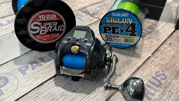 A black and silver casting fishing reel with blue line spool, displayed alongside two spools of fishing line on a printed wood surface. One spool shows 'Yo-Zuri Super Braid' and the other 'Sunline Siglon PEx4'.