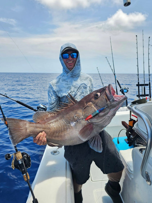 A man wearing a light blue hooded shirt and sunglasses stands on a boat holding a large fish with a colorful lure in its mouth, with fishing rods and blue ocean water in the background.