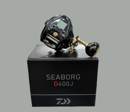 A black Daiwa Seaborg G400J fishing reel with gold and black components, displayed on top of its black product box against a solid gray background.