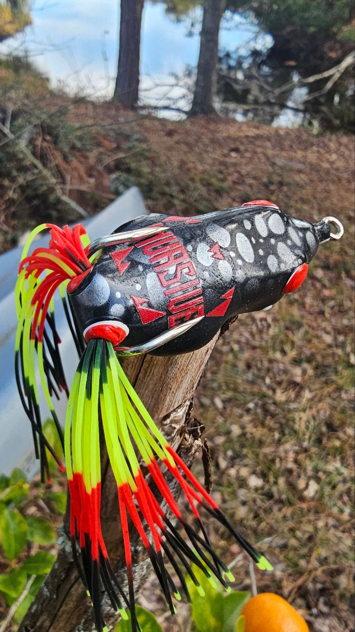 A close-up of a fishing lure attached to a branch outdoors near water, with trees and dry leaves in the background. The lure is black with red and white graphics and has bright neon yellow, red, and black skirt strands.