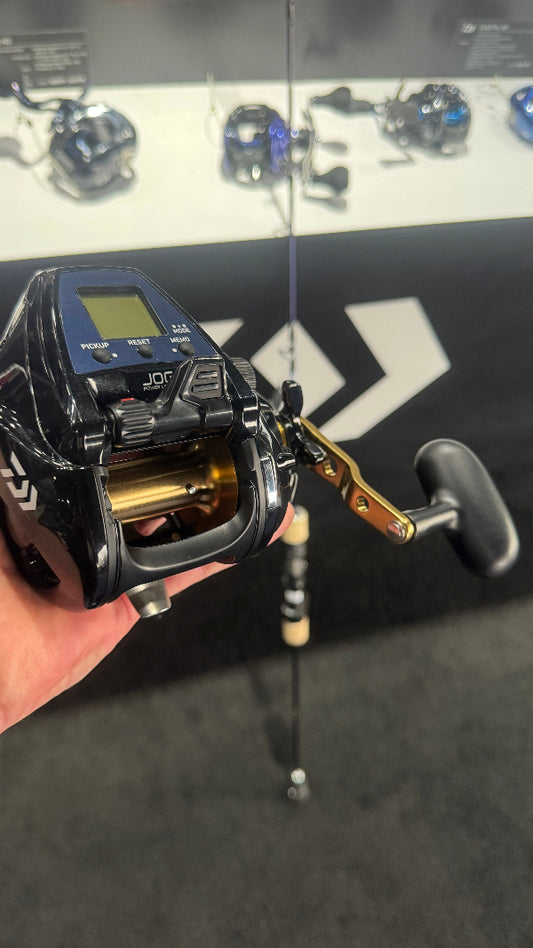 A close-up of a hand holding a black and blue Daiwa JDR fishing reel with a gold-colored spool, positioned in front of a display case containing other fishing reels.