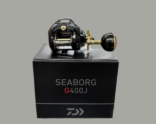 A Daiwa Seaborg G400J baitcasting fishing reel with a black and gold finish, displayed on top of its black product box against a solid gray background.