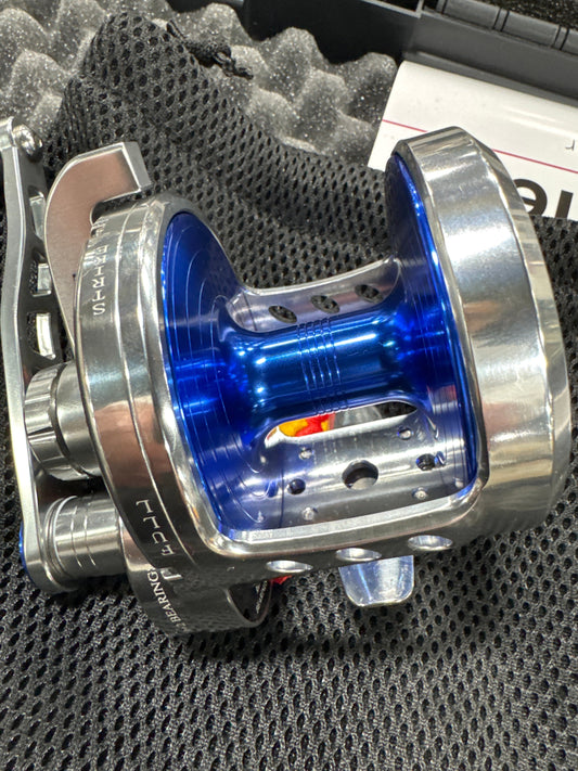 A close-up of a metallic spinning fishing reel with a polished silver body and a vibrant blue inner spool, resting on a textured black mesh surface.