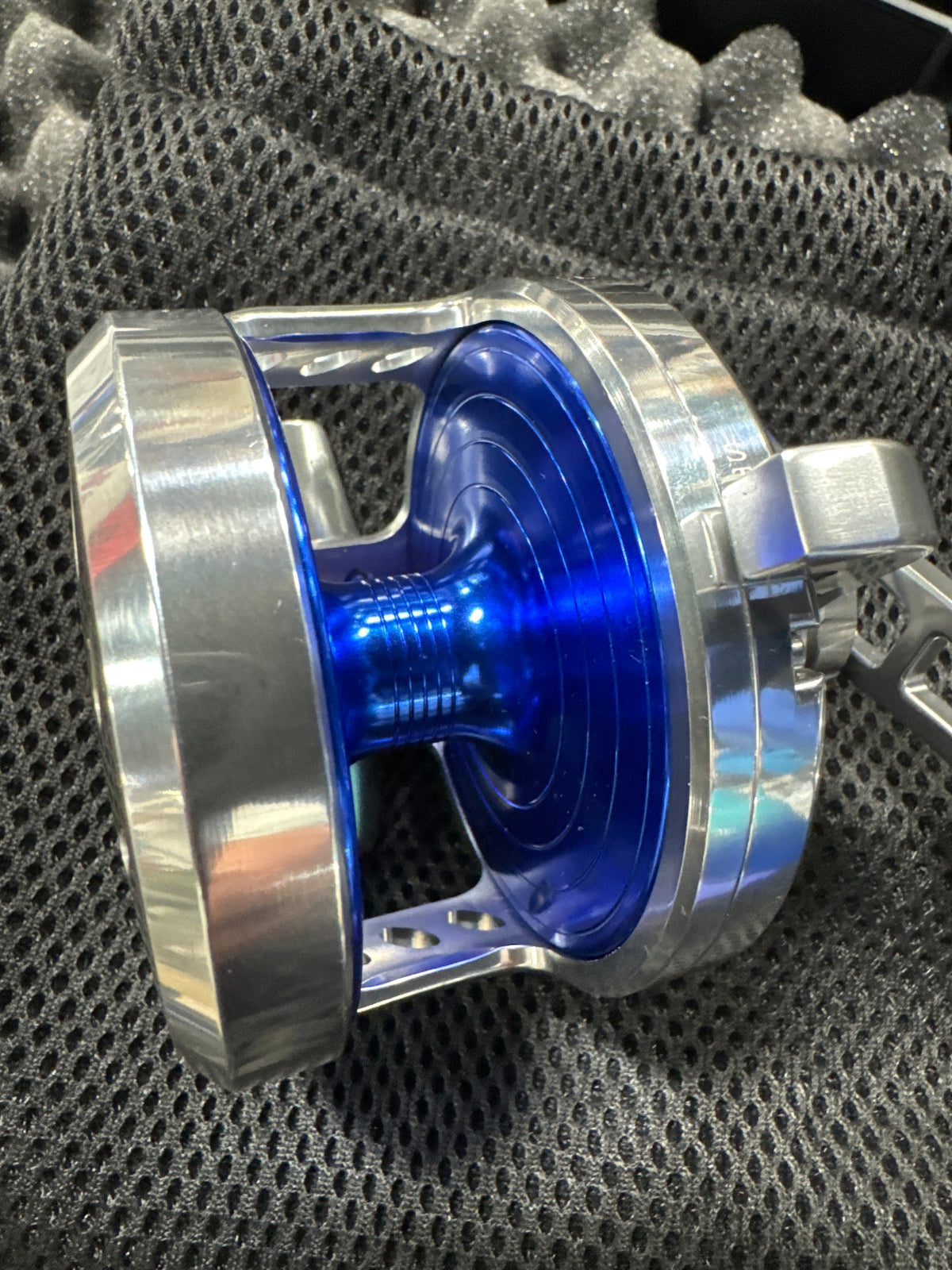 A close-up of a metallic silver and blue spool or reel component resting on a dark gray mesh fabric background.