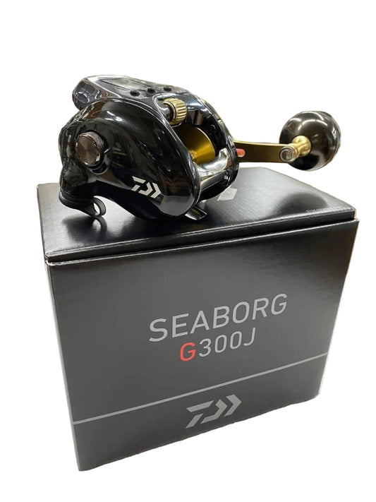 A Daiwa Seaborg G300J fishing reel with a glossy black body, gold accents, and a black handle, displayed on top of its black product box against a white background.
