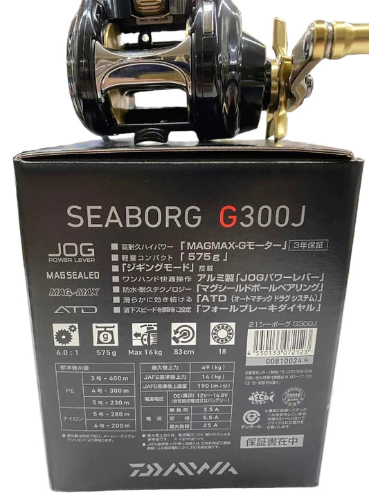 Seaborg G300J fishing reel on top of its black product packaging box, showing the back of the box with specifications and the reel's metallic black and gold components.