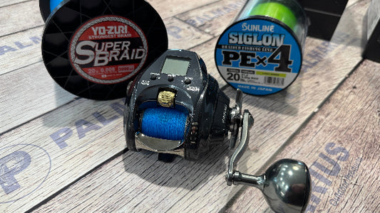 A black baitcasting fishing reel with blue line spool, shown with two spools of fishing line on a light wood surface. The reel has a silver handle and a small digital display.