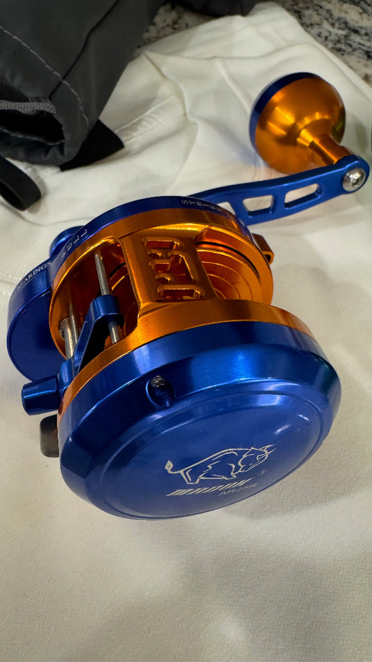 A close-up of a blue and orange fishing reel resting on a white fabric surface, with part of a black bag visible in the upper left corner.